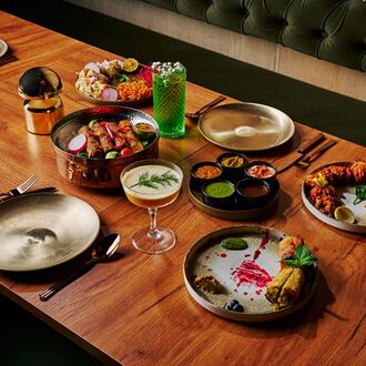 A group of several plates of indian food with colorful cocktails and gold place settings on a wooden table