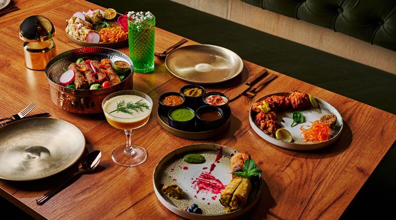 A group of several plates of indian food with colorful cocktails and gold place settings on a wooden table