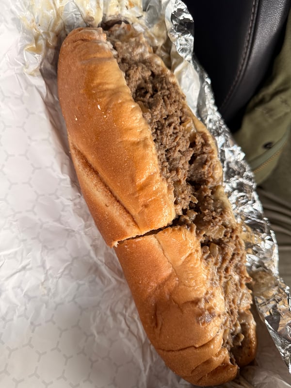 A stuffed cheesesteak sandwich cut in half and cradled in an aluminum foil wrapper is presented to the camera
