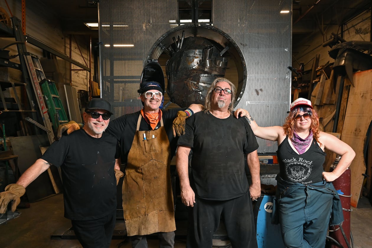 Charlie “Blackcat” Smith (third from left) poses with other artists who've worked on "The Burning Book" sculpture for Burning Man: (from left) Michael Marlingde Cuellar, Jeffery “PAPR” Morgan and Cloe Ashton. The sculpture (seen in the background) will appear at the annual Burning Man festival in Nevada’s Black Rock Desert. (Hyosub Shin/AJC)