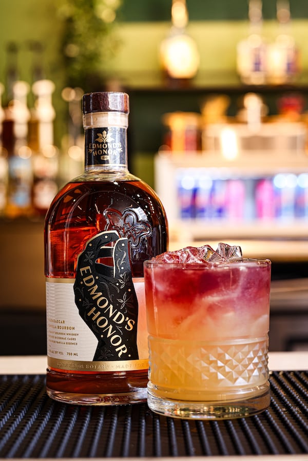 An ombre cocktail in a rocks glass full of ice. The top of the drink is a vibrant pink and the bottom is a pale orange. Next to the drink is a full bottle of Edmund's Honor bourbon. The glass and the bottle are sitting on a mat placed on the bar at the Upper Room.