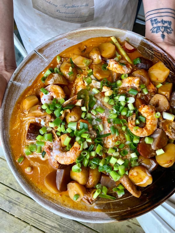 A hand presents a large skillet filled with a shrimp and potato curry, garnished with diced green onion tops.