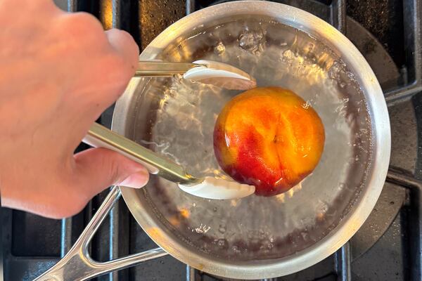 A hand hold a pair of tongs that hold a peach in boiling water.