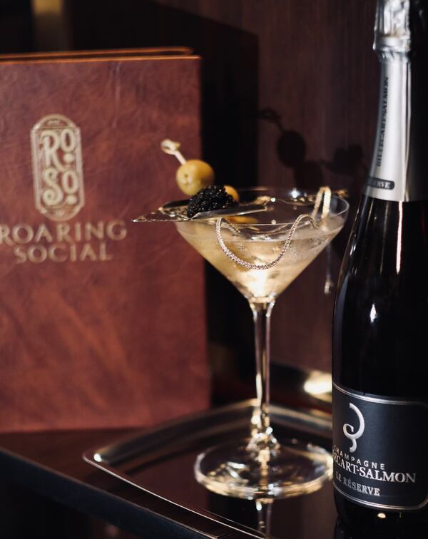A martini is garnished with olives, a spoonful of caviar and a diamond tennis necklace. It sits next to a bottle of Champagne and menu from Roaring Social.