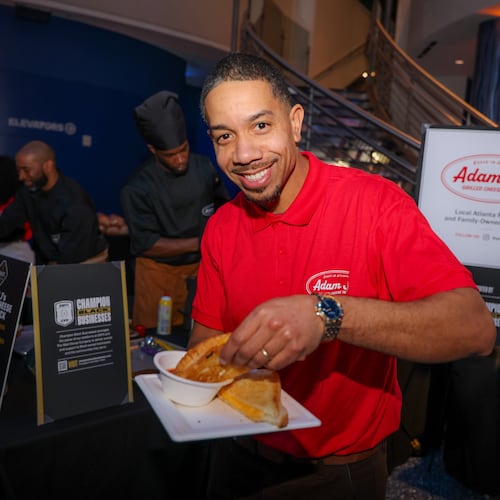 Adam Jackson is smiling and holding up a grilled cheese half. He is wearing a polo shirt with his pop-up logo and is standing in front of a sign with his logo.