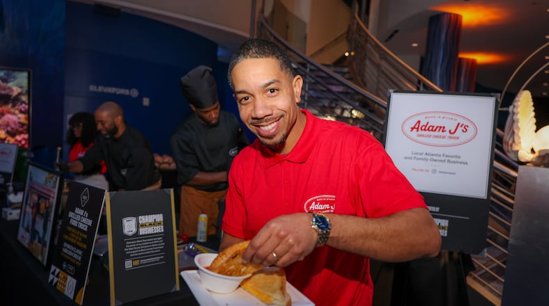Adam Jackson is smiling and holding up a grilled cheese half. He is wearing a polo shirt with his pop-up logo and is standing in front of a sign with his logo.