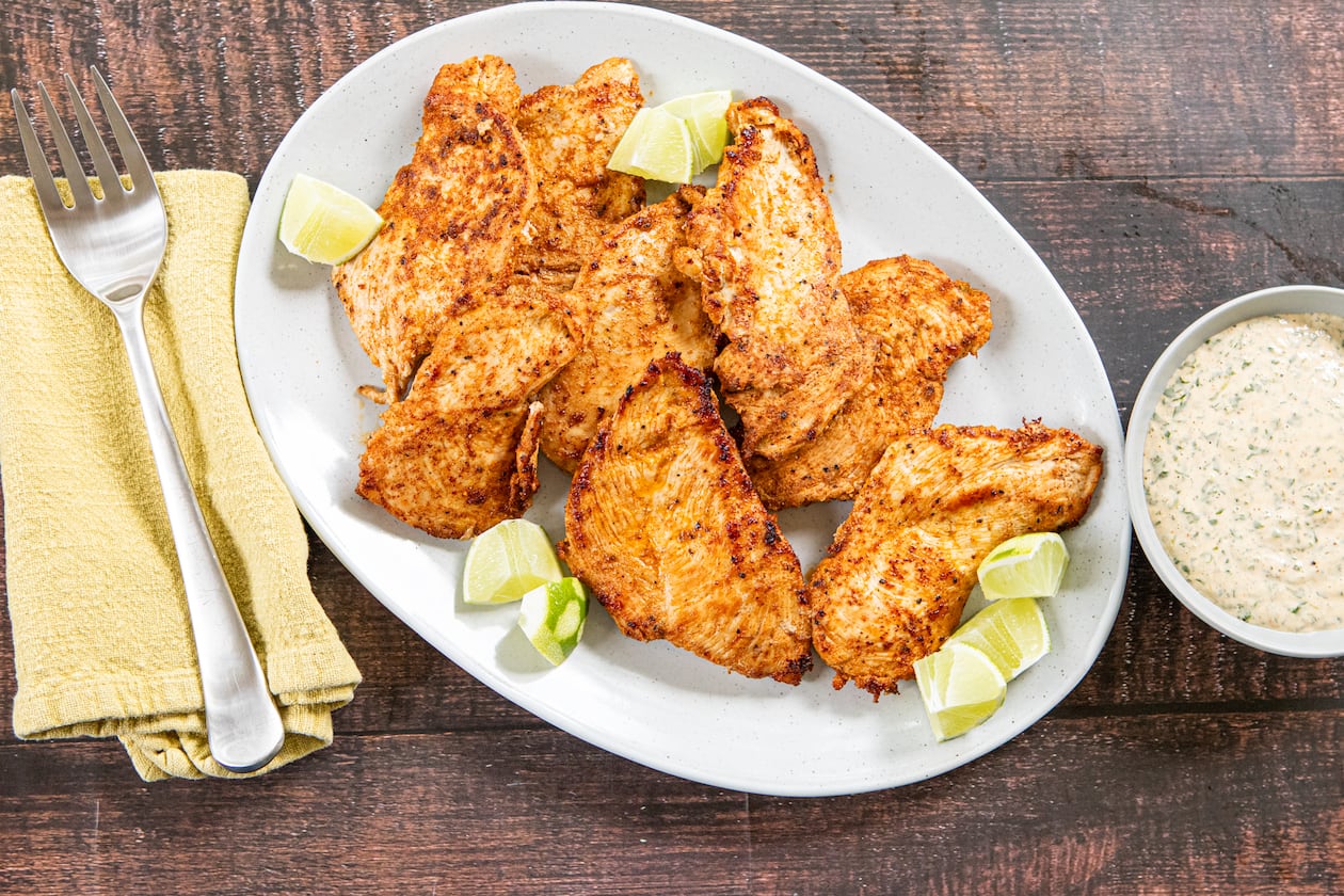 A serving plate of chicken breasts garnished with lime wedges is shown on a wood-topped table. The plate has a serving fork on a napkin next to it, as well as a bowl of dipping sauce on the other side.
