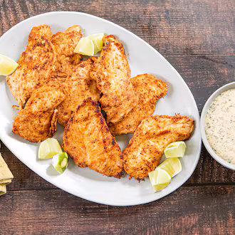 A serving plate of chicken breasts garnished with lime wedges is shown on a wood-topped table. The plate has a serving fork on a napkin next to it, as well as a bowl of dipping sauce on the other side.
