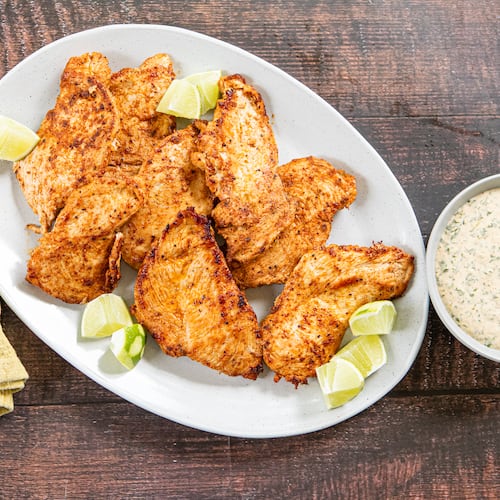 A serving plate of chicken breasts garnished with lime wedges is shown on a wood-topped table. The plate has a serving fork on a napkin next to it, as well as a bowl of dipping sauce on the other side.
