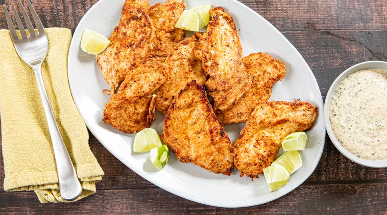 A serving plate of chicken breasts garnished with lime wedges is shown on a wood-topped table. The plate has a serving fork on a napkin next to it, as well as a bowl of dipping sauce on the other side.