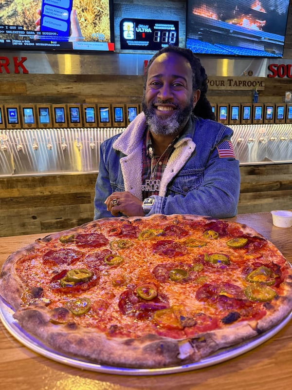 A man in a denim jacket sits behind a table holding a large pizza.