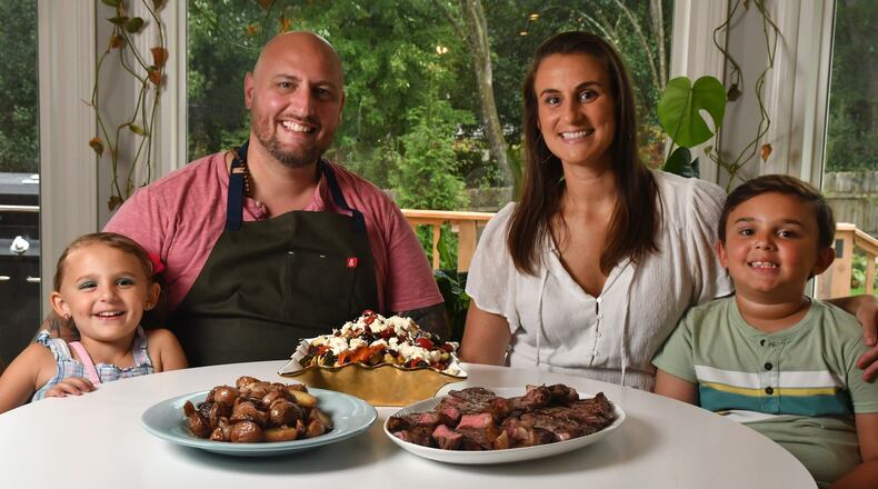 Chef Pat Pascarella sits with his wife, Megan, daughter Sophia, 5, and son Colin, 7, in the family's newly purchased Roswell home. The family is shown with one of Pascarella's favorite at-home dishes, New York Strip Steaks with Ciambotta Salad. There's also a bowl of grilled potatoes with onions because the kids want carbs, he says. (Styling by Pat Pascarella / Chris Hunt for the AJC)