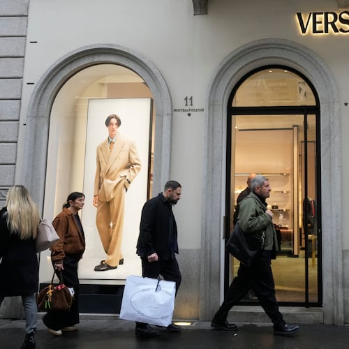 A man walks with a Prada shopping bag in front of a Versace shop, at the Montenapoleone luxury fashion street, in Milan, Italy, Tuesday, Dec. 2, 2025. (AP Photo/Luca Bruno)