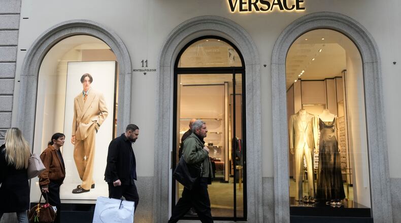 A man walks with a Prada shopping bag in front of a Versace shop, at the Montenapoleone luxury fashion street, in Milan, Italy, Tuesday, Dec. 2, 2025. (AP Photo/Luca Bruno)