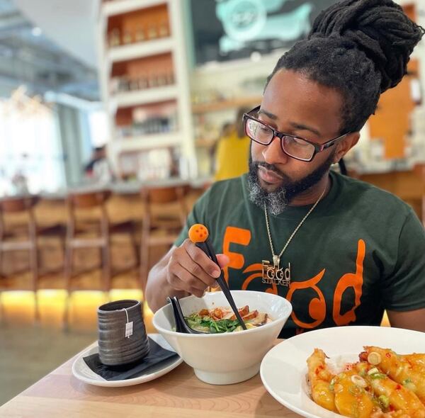 Justin Brown sits at a table at E Ramen + holding a set of chopsticks in his bowl of ramen. A mug of tea and a dish of shrimp saku-saku (fried shrimp) are also on the table.