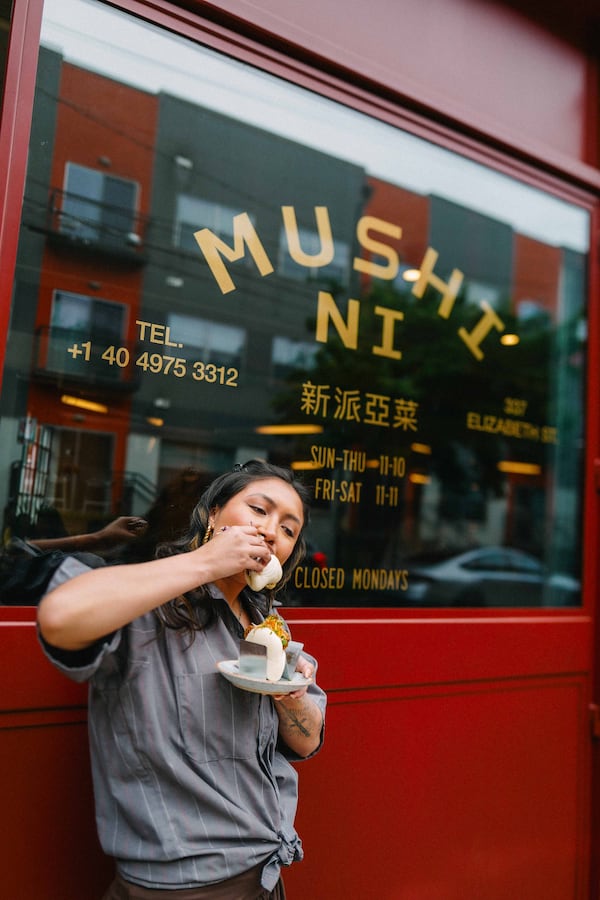 A woman eats a bao bun in front of the restaurant, which has the name Mushi Ni stenciled on the front window above her head.