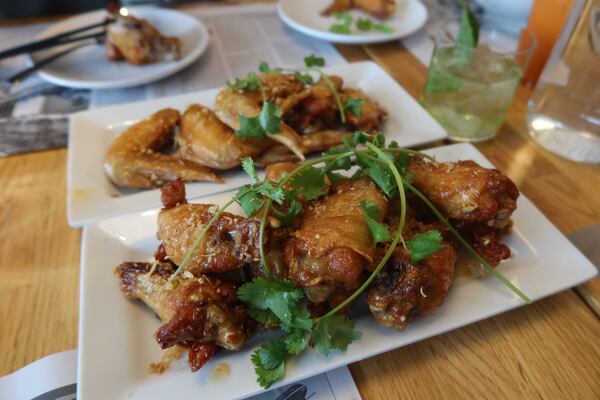 Two orders of chicken wings garnished with fresh cilantro are on plates on a restaurant table. Also in the photo is a beverage in a short glass and an open magazine, as if someone is reading the magazine while eating wings and enjoying a cocktail.