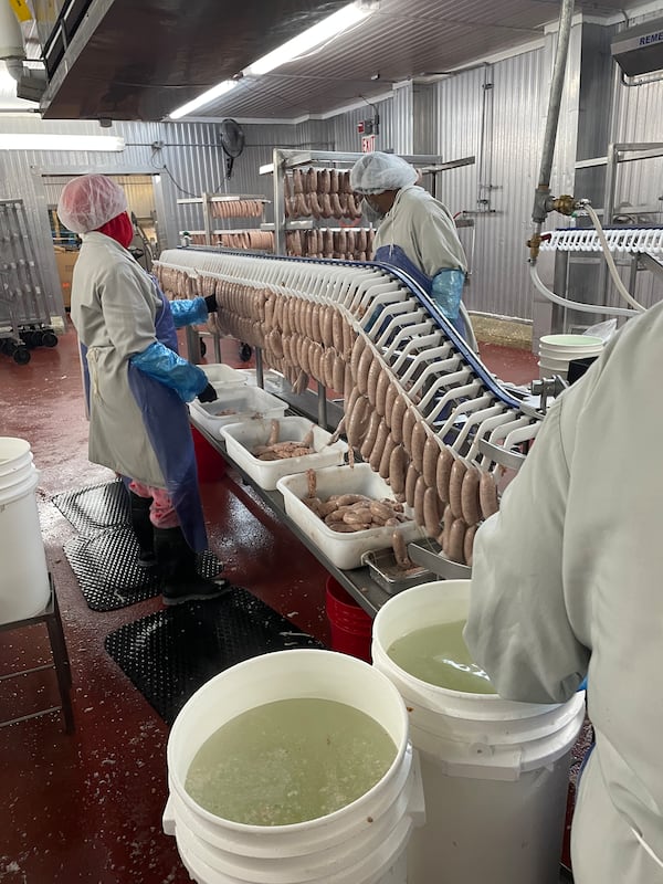 Factory workers in food safety gear handle link sausages hanging from a machinery line