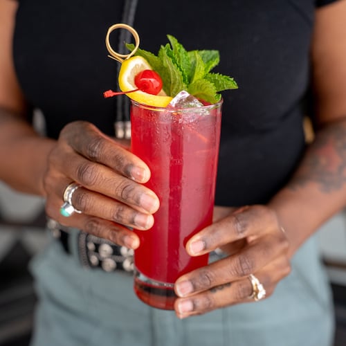 Two hands presenting a bright, cheerful-looking cocktail in a highball glass, garnished with an orange slice, cherry and a mint sprig.