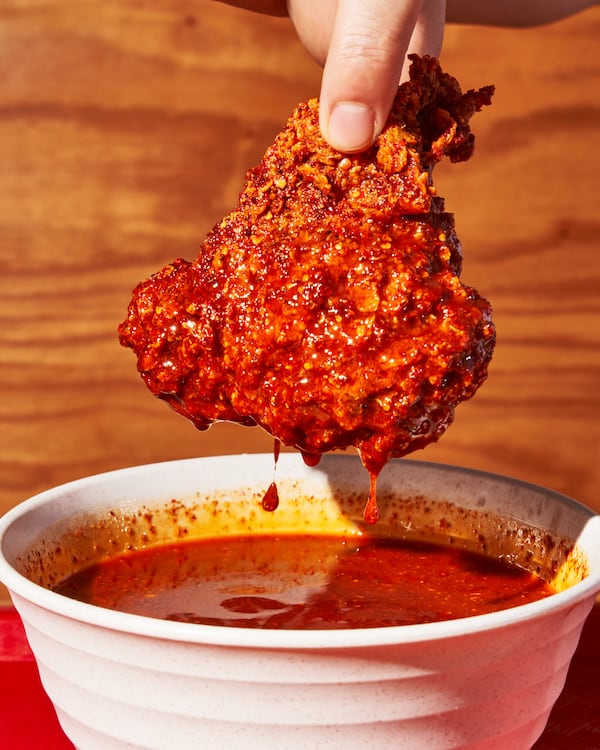 A hand is dipping a large piece of fried chicken into a plastic container of a spicy red hot sauce. The sauce is dripping off the chicken.