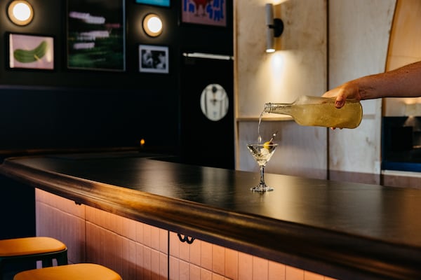 An outstretched arm reaches into the frame from the right side, pouring a martini from a large bottle into a martini glass.