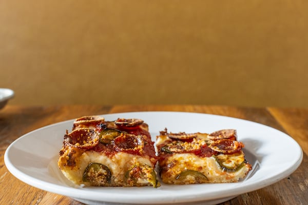 A piece of Detroit-style pizza with peppers and pepperoni sits on a plate next to a piece of skinny square pizza from Emmy Squared, showing that the original crust on the left is about twice as tall as the skinny square.