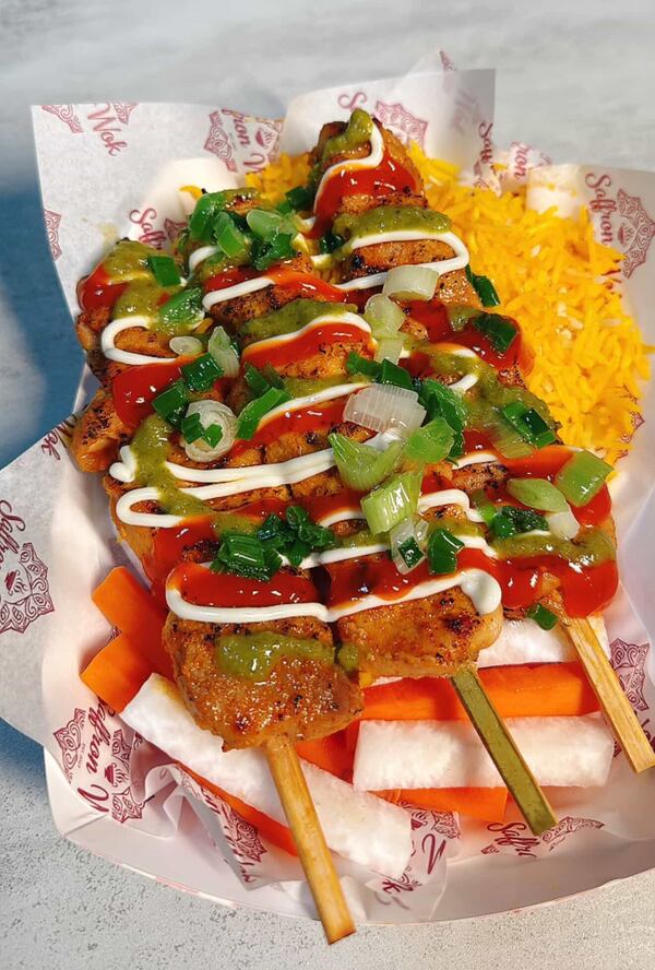 Three kabobs are lined up in a paper boat dish. They are drizzled with sauces and garnished with green onions and peppers. Also in the boat are cut vegetables and saffron rice.