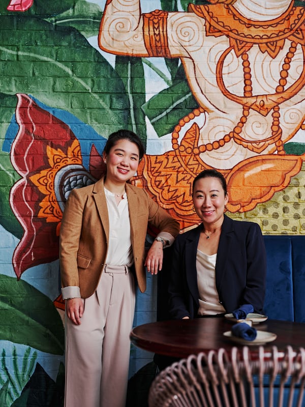 Two women pose for a photo, the woman on the left standing and the woman on the right sitting.