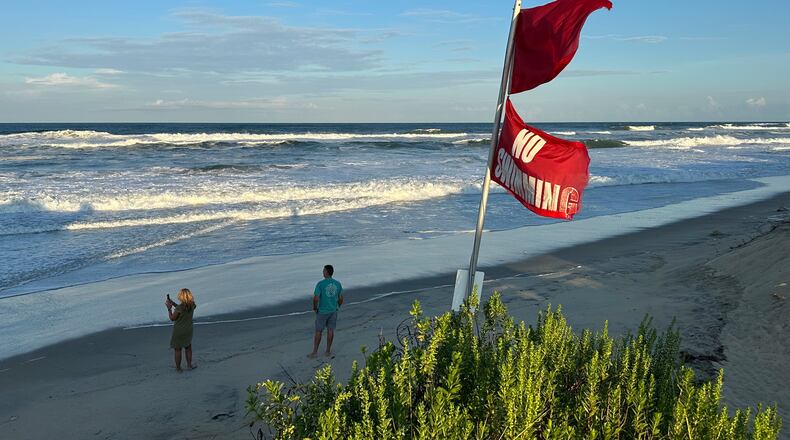 As waves from Hurricane Erin crash and “No Swimming” flags flap in the breeze, people walk on the beach in Nags Head, N.C., on Tuesday, Aug. 19, 2025. (AP Photo/Allen G. Breed)