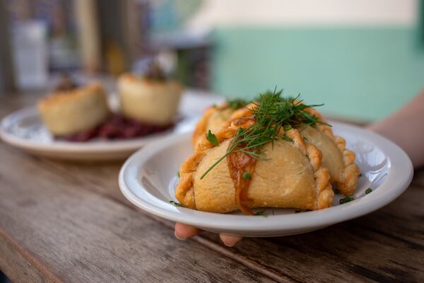 A hand extends a plate of 3 pyzys garnished with a fresh herb toward the camera. A second dish holding what might be two souffles (but is not discernible) is out of focus in the background.