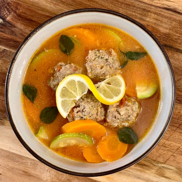 A bowl of este plato de sopa de albondigas, a meatball soup, containing meatballs, vegetables, fresh herb leaves and garnished with a lemon twist.
