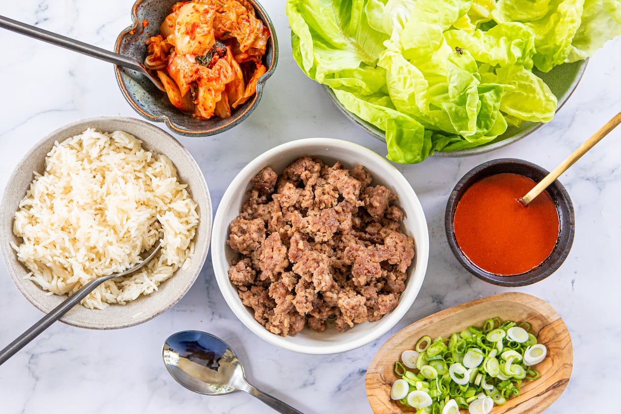A collection of prepared foods arranged in serving dishes for a family-style meal. Shown are bowls containing white rice, browned ground pork, a homemade gochujang sauce, chopped green onions, lettuce leaves and kimchi. Spoons are tucked into or next to some of the bowls.