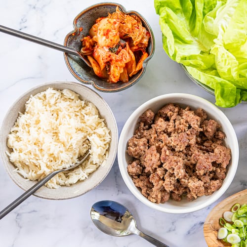 A collection of prepared foods arranged in serving dishes for a family-style meal. Shown are bowls containing white rice, browned ground pork, a homemade gochujang sauce, chopped green onions, lettuce leaves and kimchi. Spoons are tucked into or next to some of the bowls.