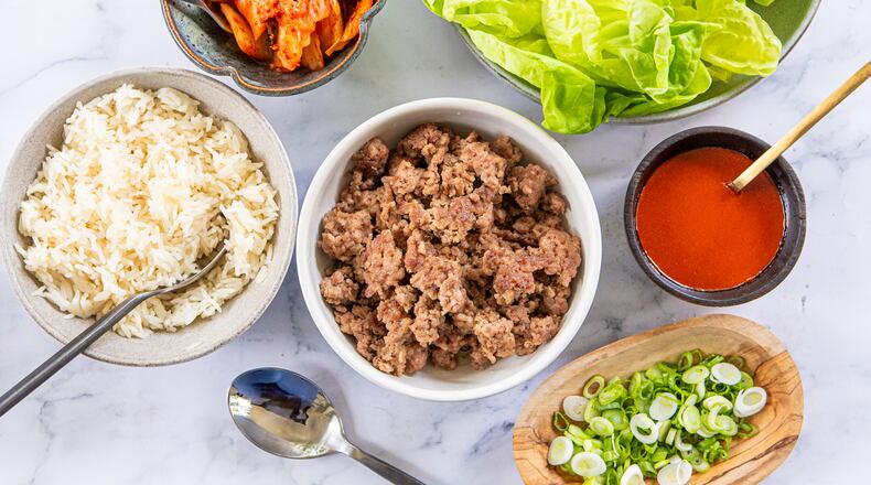 A collection of prepared foods arranged in serving dishes for a family-style meal. Shown are bowls containing white rice, browned ground pork, a homemade gochujang sauce, chopped green onions, lettuce leaves and kimchi. Spoons are tucked into or next to some of the bowls.