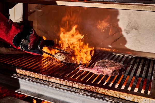 A gloved hand holding tongs handles a steak on a stainless steel grill with flames leaping around it
