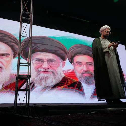 A cleric checks his cell phone on stage in front of a screen displaying portraits of the late Iranian revolutionary founder Ayatollah Khomeini, left, late Supreme Leader Ayatollah Ali Khamenei, center, and Supreme Leader Ayatollah Mojtaba Khamenei, during a pro-government gathering in Tehran, Iran, Sunday, June 7, 2026. (AP Photo/Vahid Salemi)