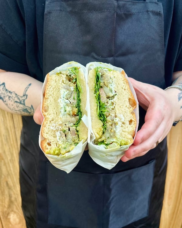 A person wearing an apron holds a sandwich cut in half in front of their midsection, showing the cross section of the porchetta sandwich.
