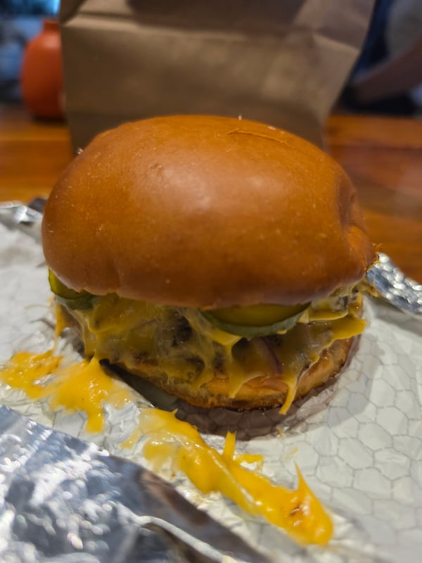 A cheeseburger with generous amounts of cheese, both dripping off the burger and in a wide line on the aluminum foil wrapper. The burger has a plain bun and has several visible pickle slices peeking out. A brown paper bag is in the background.