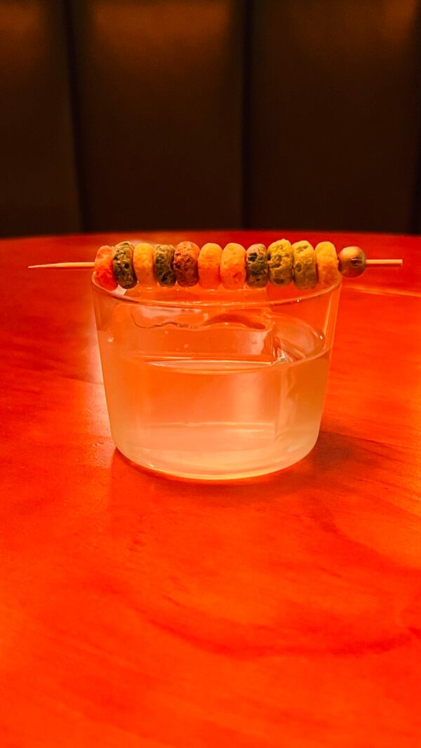A color-saturated  photo of a clear cocktail in a rocks glass garnished with Froot Loops threaded on a long toothpick.