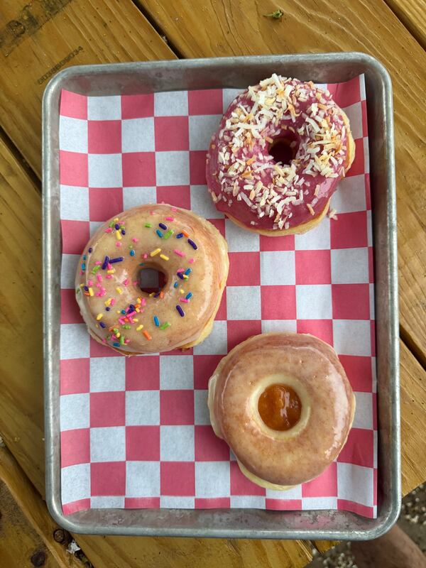 A top-down view of three gourmet donuts on a pink and white checkered paper tray liner, featuring a red-glazed donut with toasted coconut, a vanilla-glazed donut with rainbow sprinkles, and a glazed donut with peach jam.