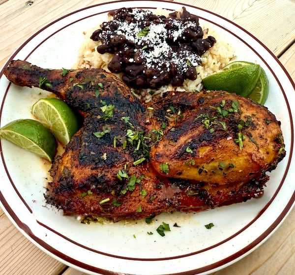 A half of grilled chicken is on a diner-style plate. It is served with Cuban beans and rice and lemon wedges.