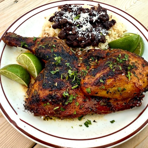 A half of grilled chicken is on a diner-style plate. It is served with Cuban beans and rice and lemon wedges.