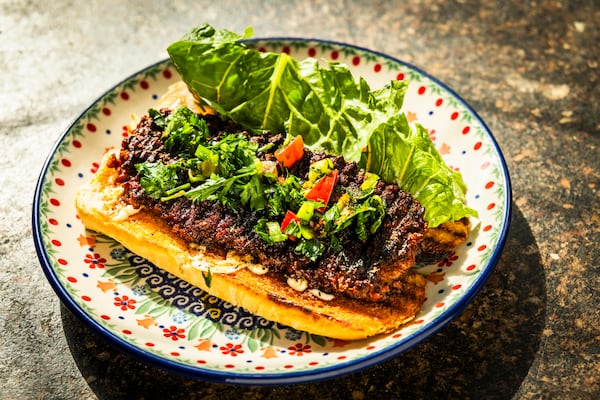 A colorful plate holds an open-faced sandwich. The bread is a toasted baguette with grill marks. There is a cream sauce drizzled on the bottom baguette half. A rectangular sausage patty, approximately the size of the baguette, is on top of the cream. The sausage has the leaves and stems of chopped parsley, on top, along with a few pieces of a chopped red tomato. A lettuce leaf is tucked next to the sandwich.