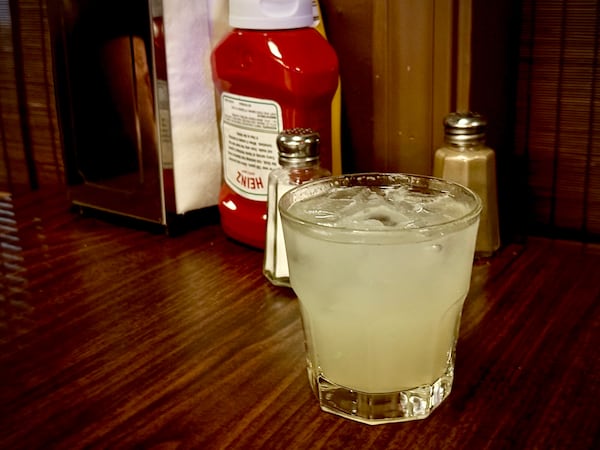 A pale yellow cocktail on a wood table in a wood-paneled room that also holds salt shakers and a Heinz ketchup bottle and a napkin dispenser