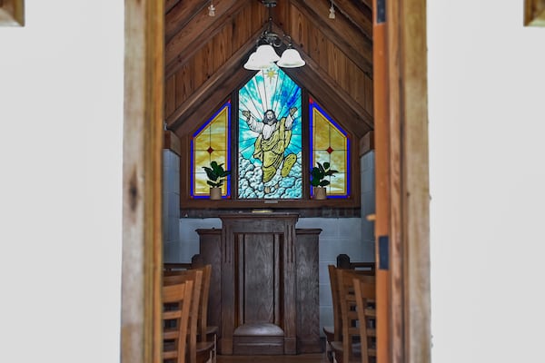 The chapel has three rows of chairs, four chairs to a row. An entrance door at one end and a pulpit in front of a stain-glassed window of Jesus at the other.  (Sarah Peacock for AJC)