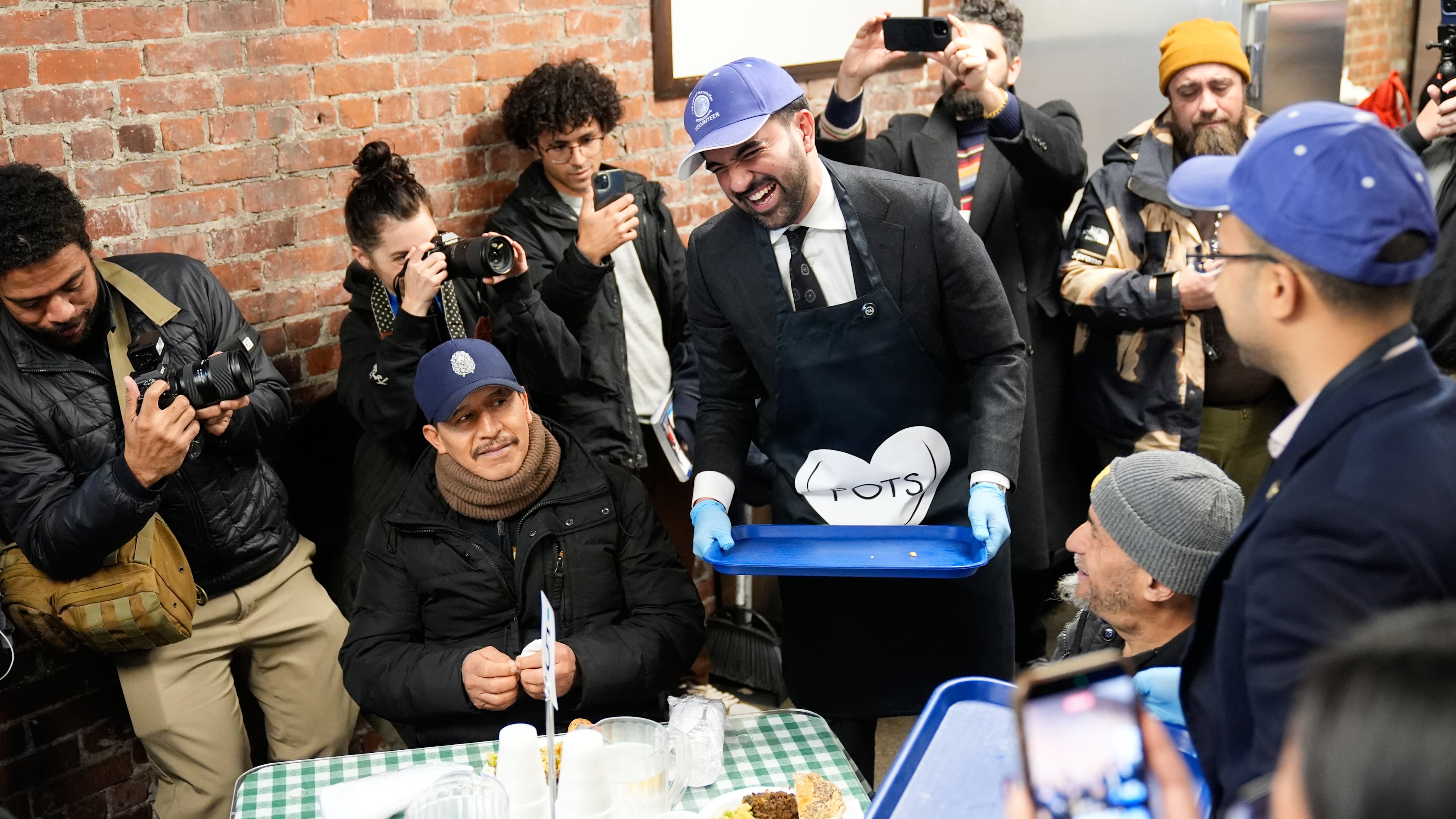 While surrounded by members of the media, New York City mayor-elect Zohran Mamdani, center, helps to serve meals at at POTS, a Bronx nonprofit that provides food assistance, in New York, Monday, Nov. 17, 2025. (AP Photo/Seth Wenig)