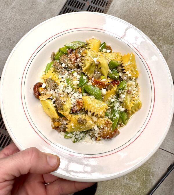 A hand is holding a bowl of pasta mixed with bright vegetables and garnished with a hefty sprinkle of cheese.