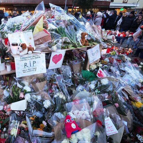 People stand around floral tributes and candles placed outside the sealed off Le Constellation bar in Crans-Montana, Swiss Alps, Switzerland, Saturday, Jan. 3, 2026, where a devastating fire left dead and injured during the New Year's celebrations. (AP Photo/ Antonio Calanni)