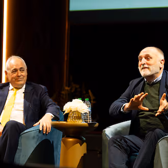 Two men sit in chairs on a stage, the man at the left smiling at the man on the right, who is talking and gesturing with his hands