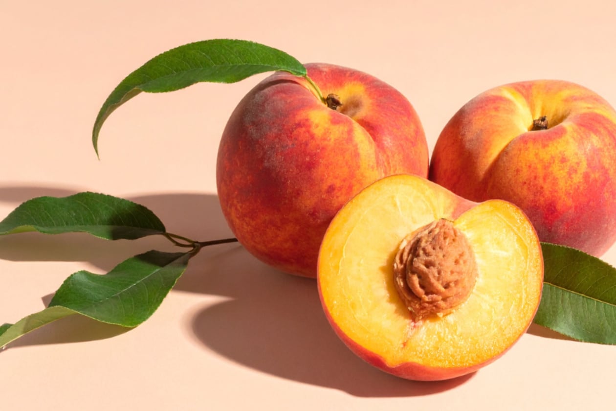 Two whole peaches sit on a peach background with peach leaves still attached to them along with a sliced peach, showcasing the seed and bright yellow flesh.